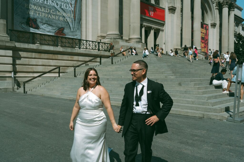 Bride and groom smile and hold hands outside of MET museum after their NYPL wedding, by Elsie Goodman.