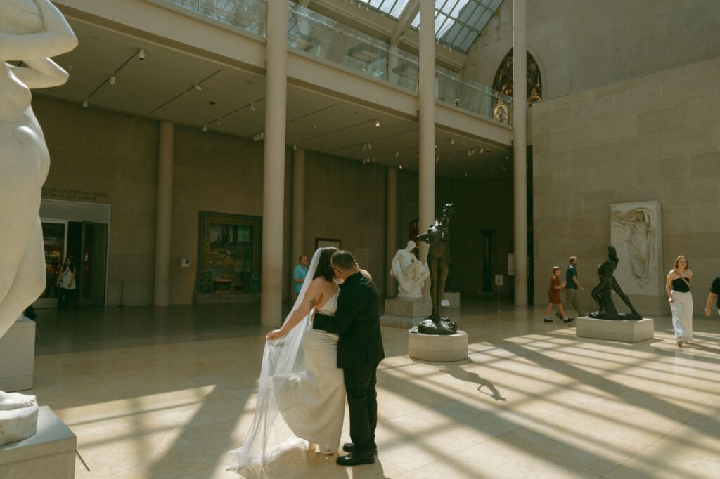 Bride and groom hug in sun spot at museum after their NYPL wedding, by Elsie Goodman.