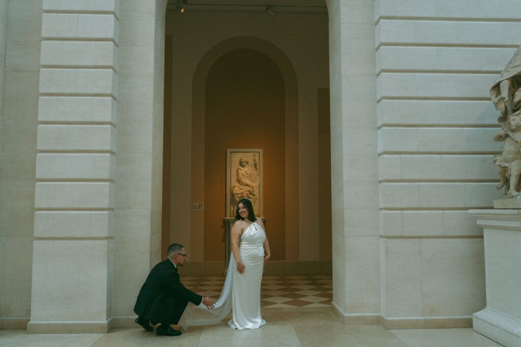 Groom fixes bride's train under arch at a museum after their NYPL wedding, by Elsie Goodman.