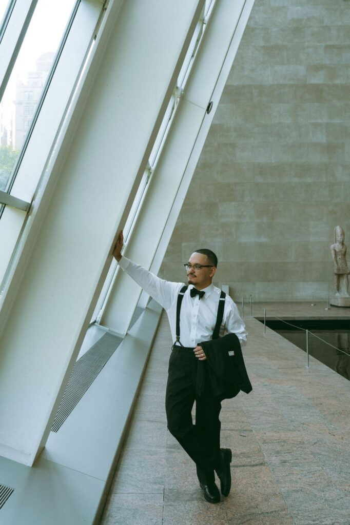 Portrait of groom leaning against sunny window with jacket over arm, captured by Elsie Goodman Photography.