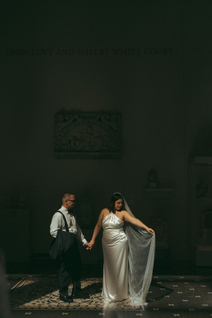 Bride fans veil while holding groom's hand against dark background, by Elsie Goodman.