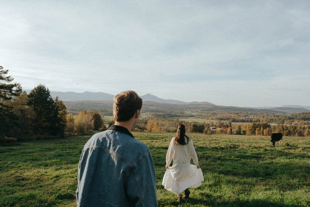 Couple walks with cows in pasture, part of their autumn in Vermont engagement photos by Elsie Goodman.