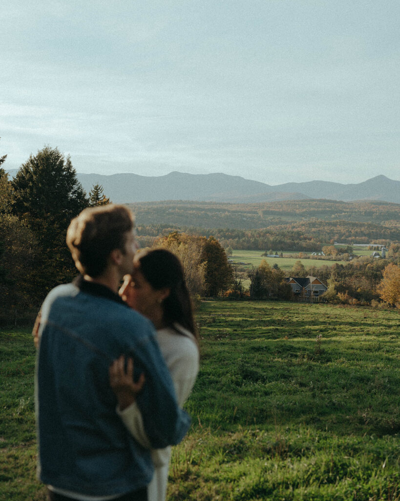 Couple hugs in soft focus in an open field during their Vermont engagement session, captured by Elsie Goodman.