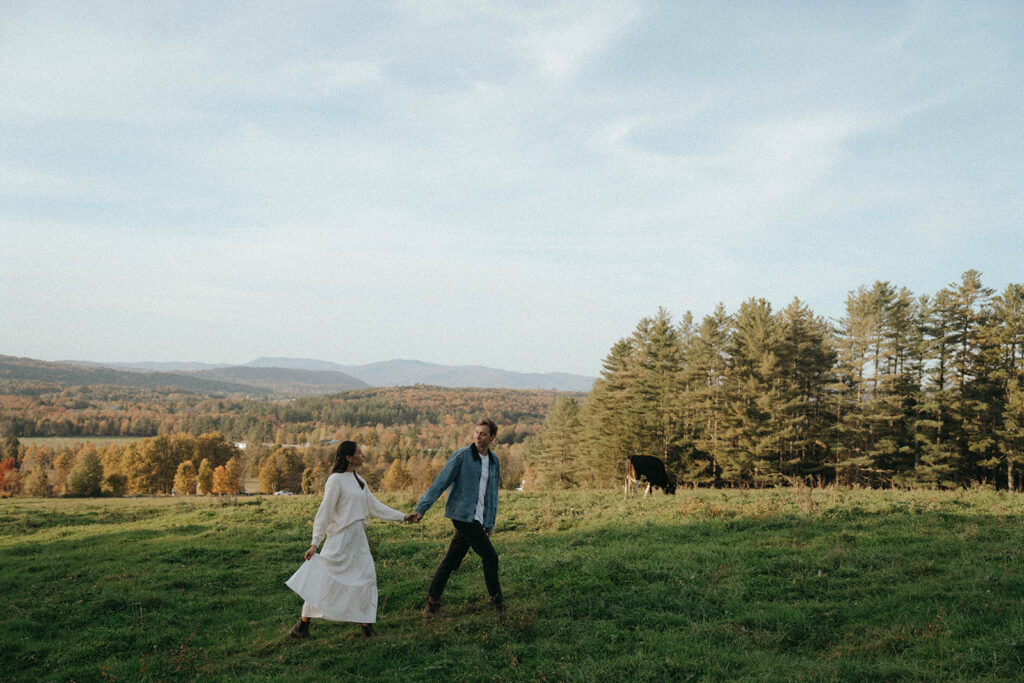 Man looks back at woman walking through pasture, with fall colors in this Vermont couples session by Elsie Goodman.