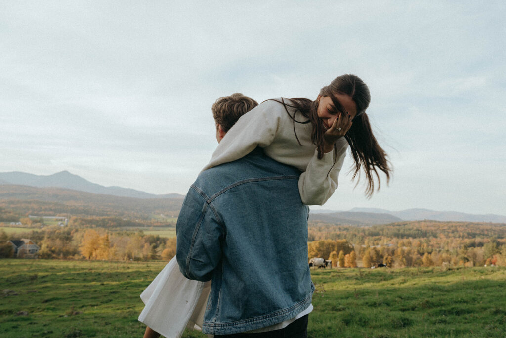 Man lifts woman playfully over his shoulder with cows and fall foliage behind, Vermont engagement by Elsie Goodman.