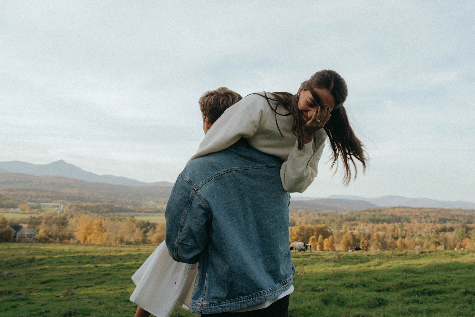 Man lifts woman playfully over his shoulder with cows and fall foliage behind, Vermont engagement by Elsie Goodman.