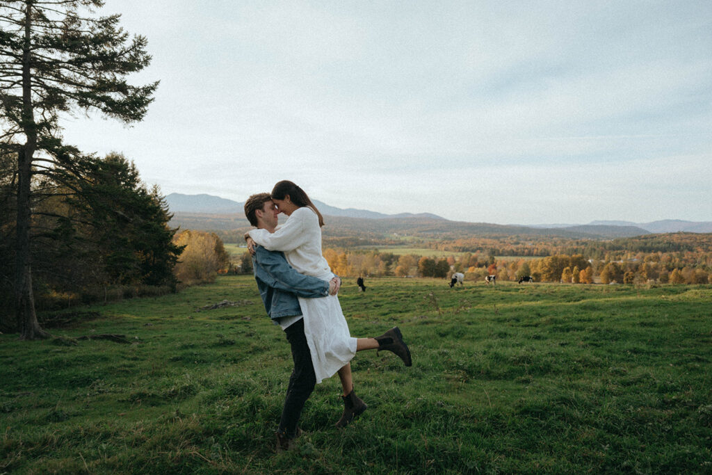 Man lifts woman in open Vermont field at sunset, surrounded by cows and fall trees, engagement photos by Elsie Goodman.