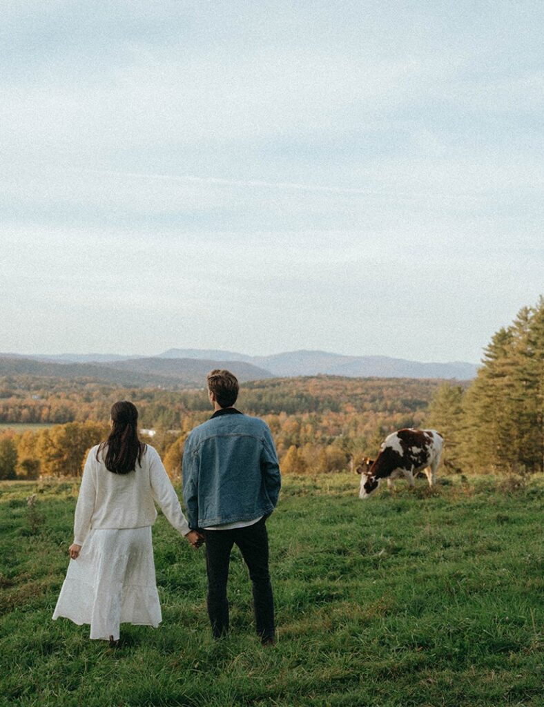 Couple stands facing Vermont mountains and fall foliage, holding hands during their engagement session with Elsie Goodman.