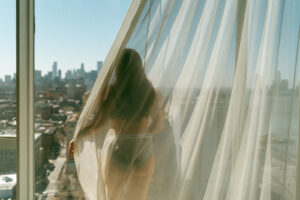 Woman in lingerie stands by a large window, sunlight streaming through sheer curtains with a city skyline view in the background, shot by Elsie Goodman Photography.