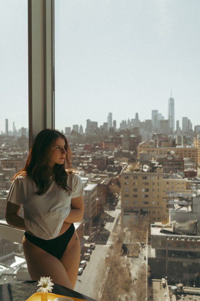 Sensual hotel boudoir shoot of woman looking out window over New York skyline, shot by Elsie Goodman Photography.