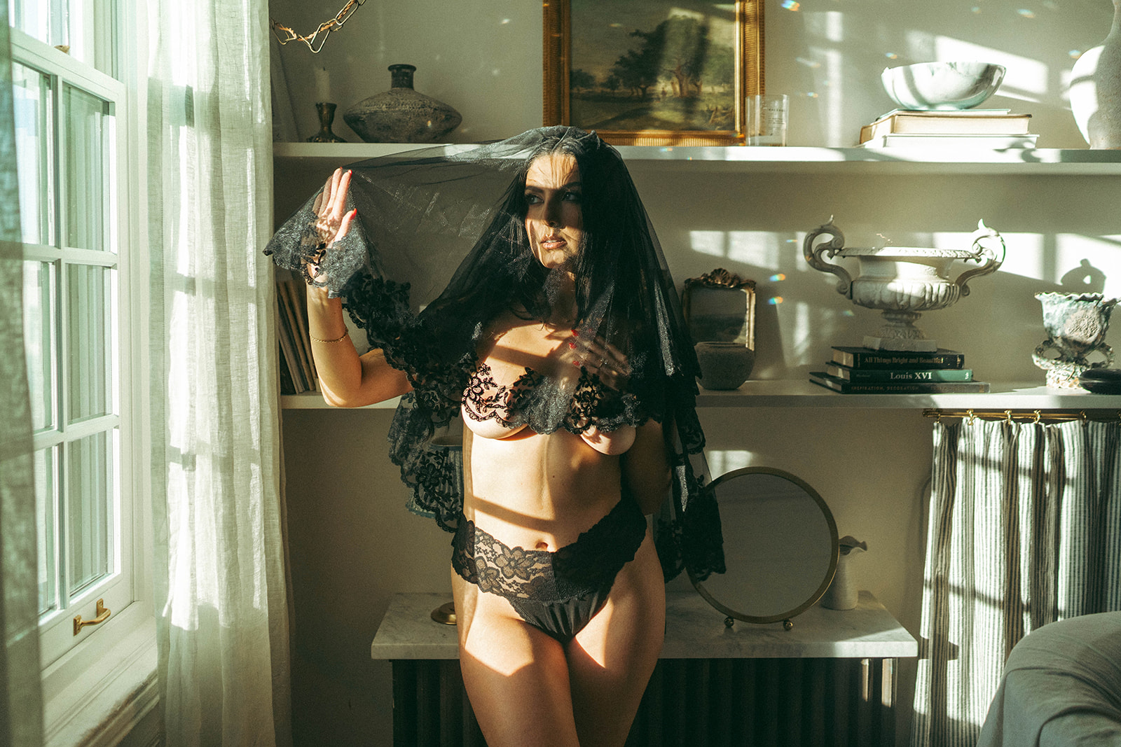 Dramatic boudoir photoshoot at home with woman lifting black lace veil while standing in sunlight, shot by Elsie Goodman Photography.