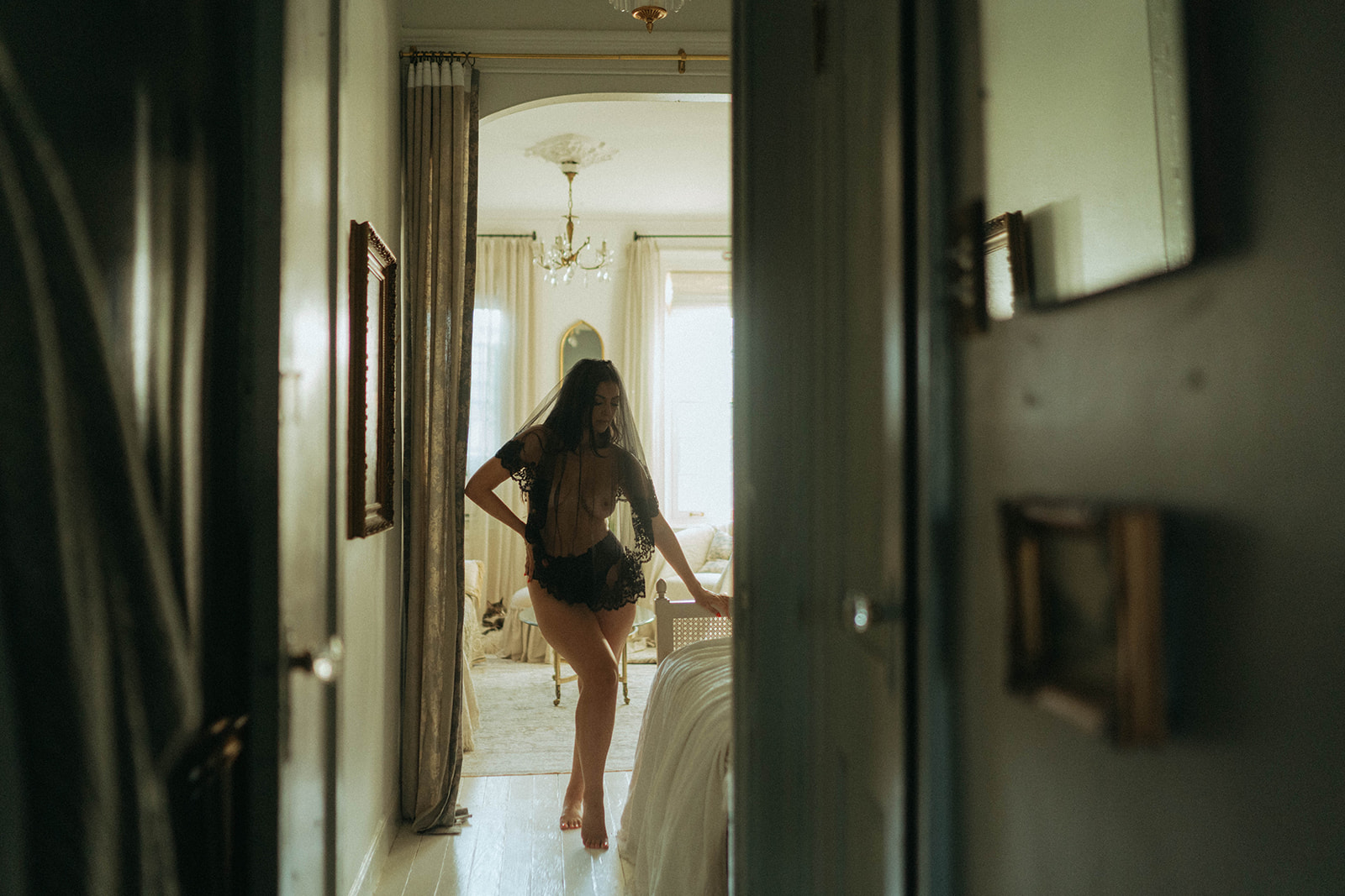 Boudoir photoshoot at home with woman walking down hallway in lingerie and veil, framed by doorway light, shot by Elsie Goodman Photography.