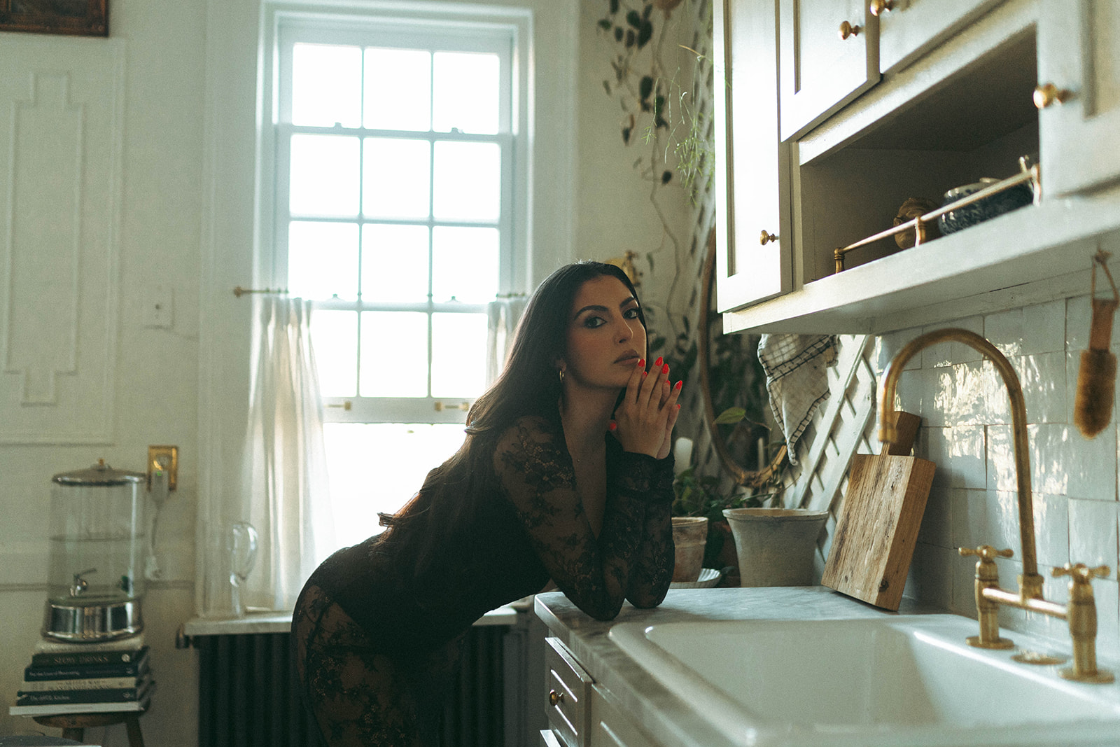 Intimate boudoir photoshoot at home featuring a woman leaning thoughtfully against a farmhouse sink, sunlight streaming through the window, shot by Elsie Goodman Photography.