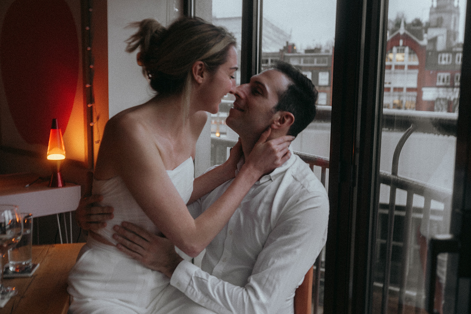 Woman sitting on a man's lap, touching noses and smiling, a dark London city street in the background, shot by London engagement photographer Elsie Goodman.