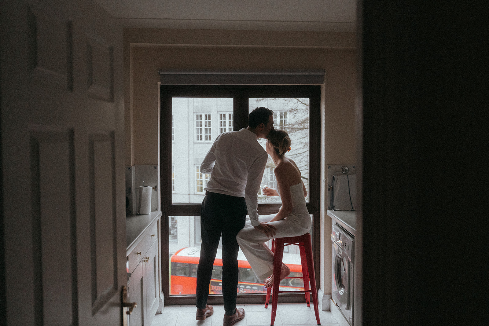 Man leaning over and giving a woman a kiss on the forehead. She is sitting on a red stool, looking out at a busy London street, shot by London engagement photographer Elsie Goodman.