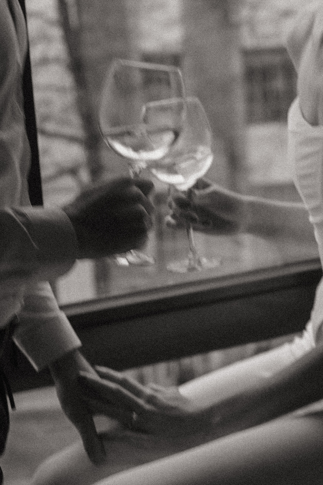 Black and white photo of a man and a woman clinking together two wine glasses. His hand is on her knee and her hand rests on top, shot by London engagement photographer Elsie Goodman.