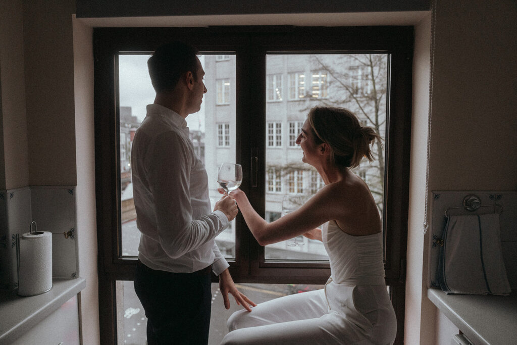 A man stands and a woman sits in front of a window that shows a grey London street, she is pointing out the window, smiling, and he is holding a wine glass, shot by London engagement photographer Elsie Goodman.