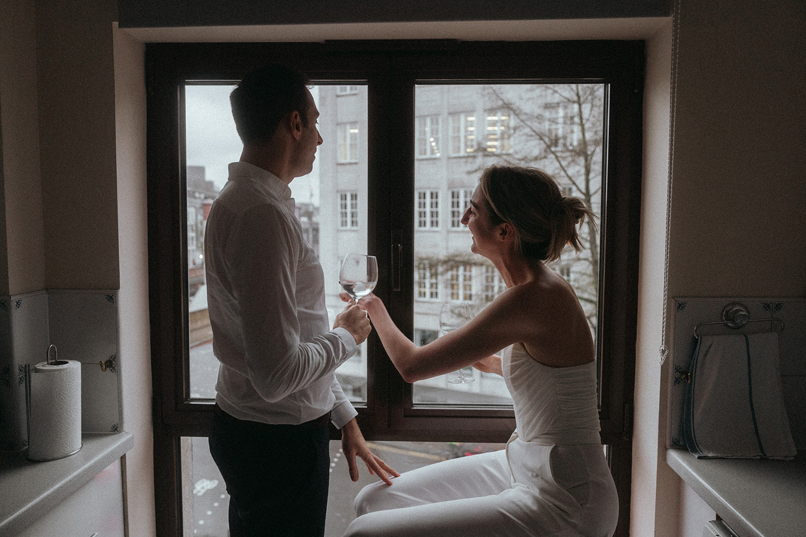 A man stands and a woman sits in front of a window that shows a grey London street, she is pointing out the window, smiling, and he is holding a wine glass, shot by London engagement photographer Elsie Goodman.