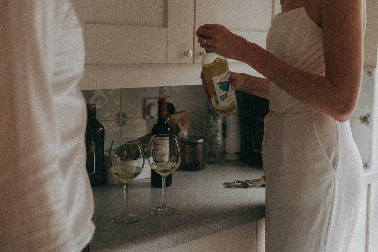 Cropped image of two wine glasses on a counter, a woman holds a bottle of white wine putting the cap back on, shot by London engagement photographer Elsie Goodman.