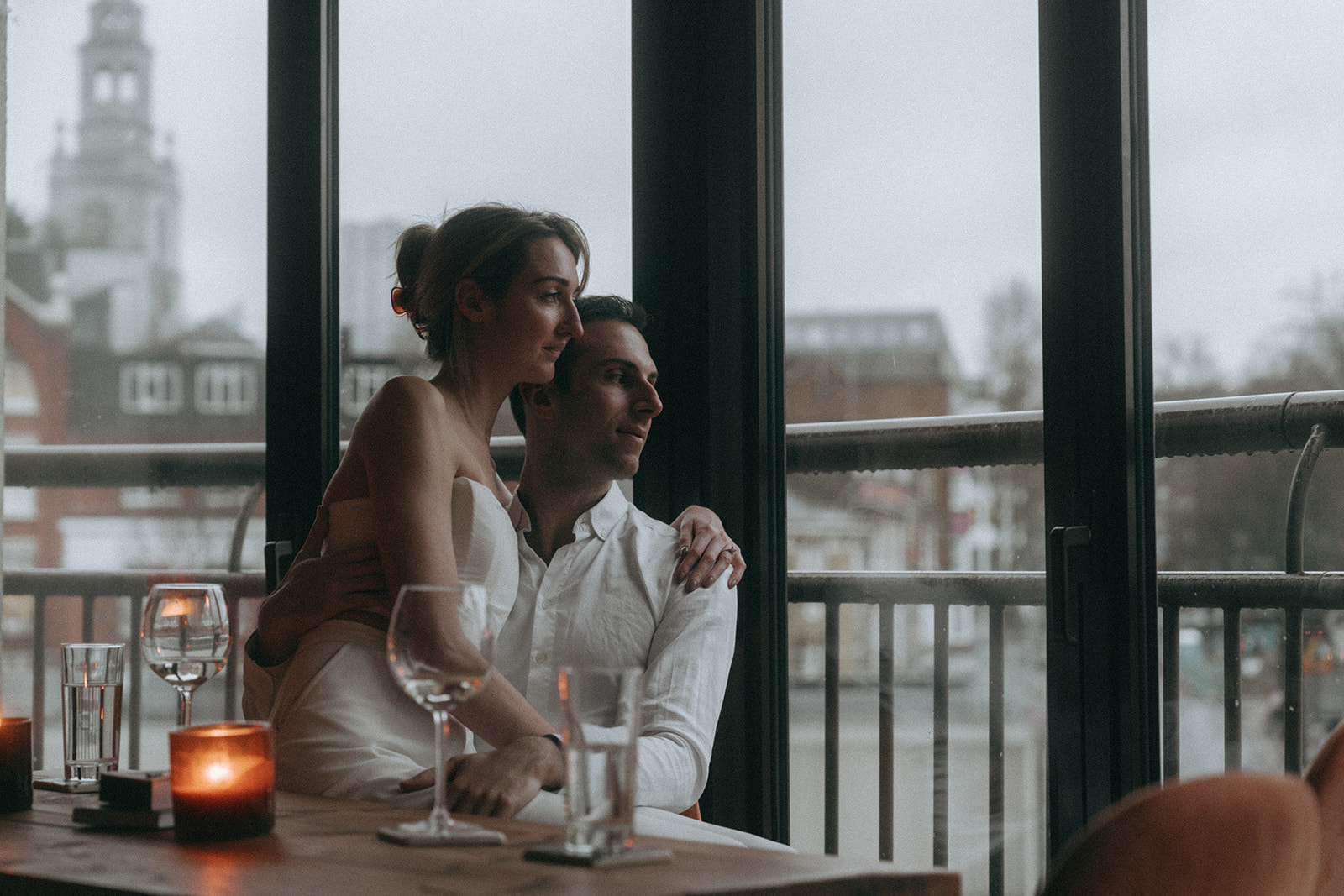 Woman sitting on a man's lap with her around him. He has his hand on her thigh, and they are both looking out the window to a rainy London scene, shot by London engagement photographer Elsie Goodman.