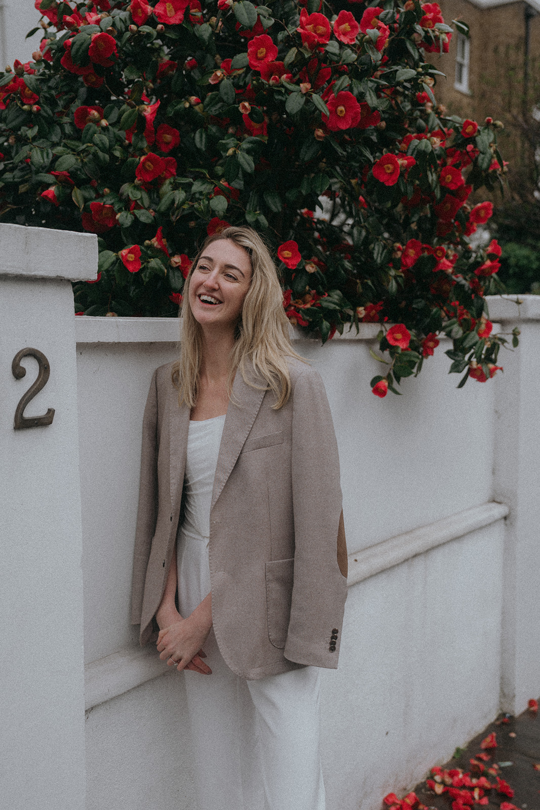 A woman leaning against a white garden wall with a big red flowery bush behind her. She has a suit jacket draped over her shoulders, smiling, shot by London engagement photographer Elsie Goodman.