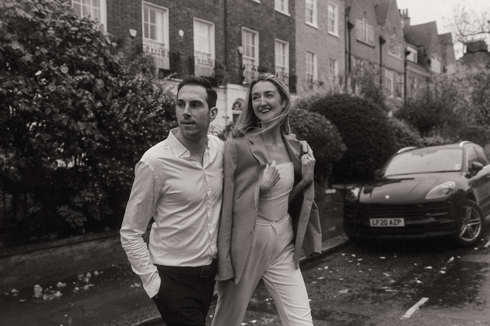 Black and white photo of a man and a women crossing a London street, she is wearing his suit jacket smiling, shot by London engagement photographer Elsie Goodman.