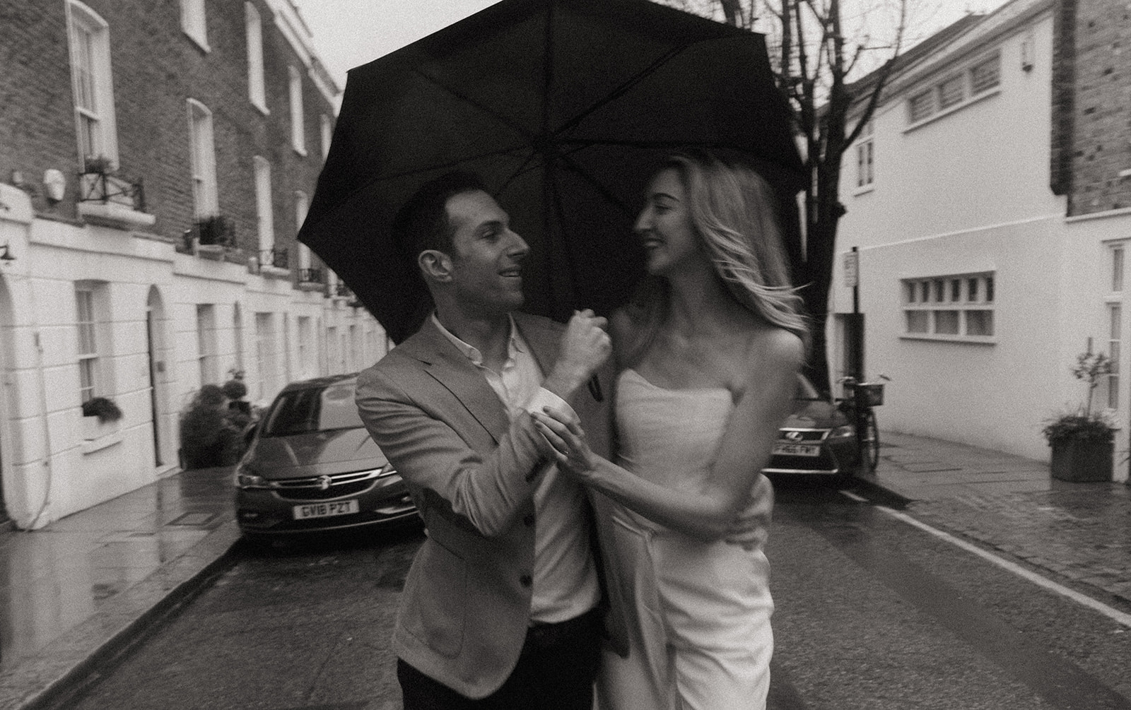 A man and a woman on a drizzly London street, he is holding an umbrella over both of them .They are both smiling, shot by London engagement photographer Elsie Goodman.