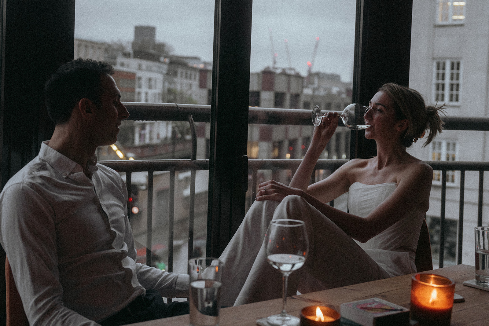 Man and women sitting at a table. She is drinking wine out of a wine glass and he is looking at her. A dark rainy London street can be seen behind them, shot by London engagement photographer Elsie Goodman.