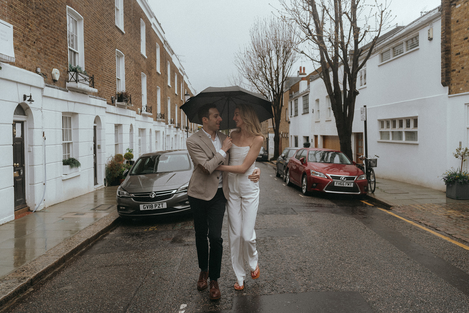 Man and women walking down a London street with an umbrella, a woman standing behind her fiancé with her arms wrapped around him lovingly on a London corner, shot by London engagement photographer Elsie Goodman.