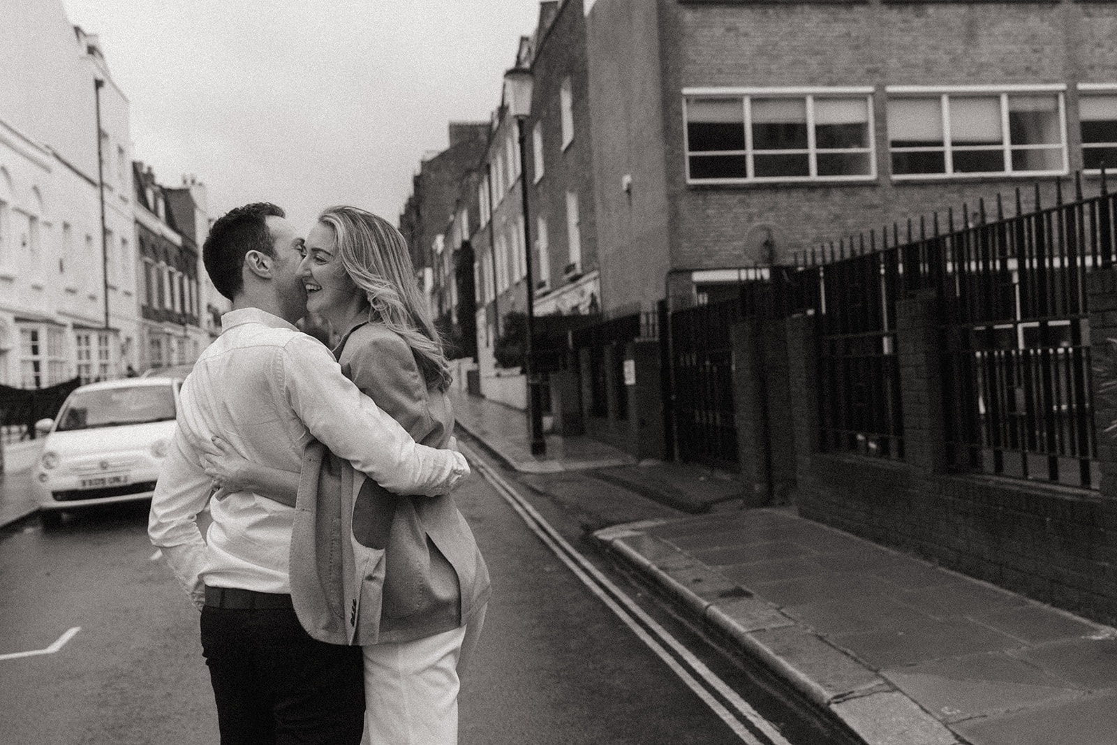 Black and white photo of a man and a woman hugging and laughing in the street, shot by London engagement photographer Elsie Goodman.