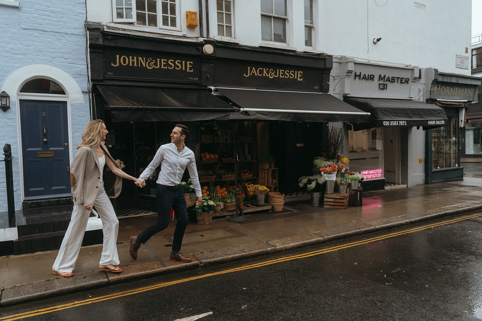 Man leading a woman down a London street by her hand. Behind them some small shops, shot by London engagement photographer Elsie Goodman.
