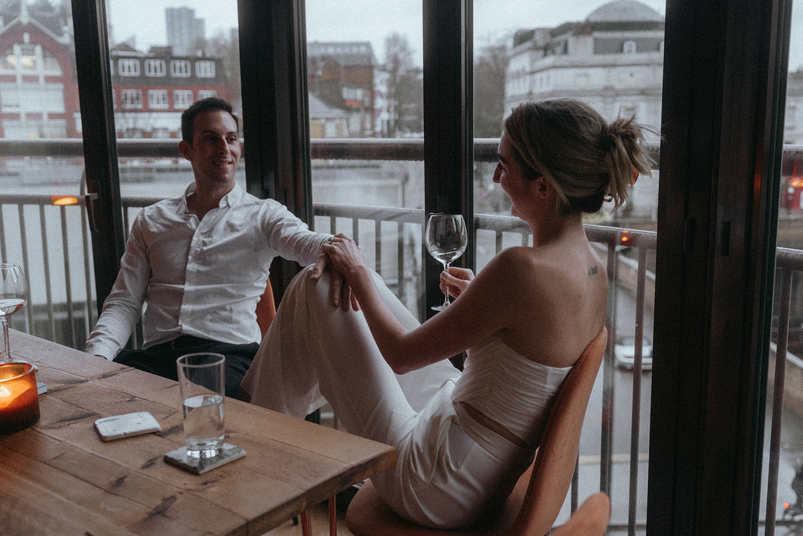 Man and women sitting at a table, a dark stormy London street can be seen in the window behind them. He has his hand on her knee, and her hand is resting on top of his; in the other is a wine glass, shot by London engagement photographer Elsie Goodman.