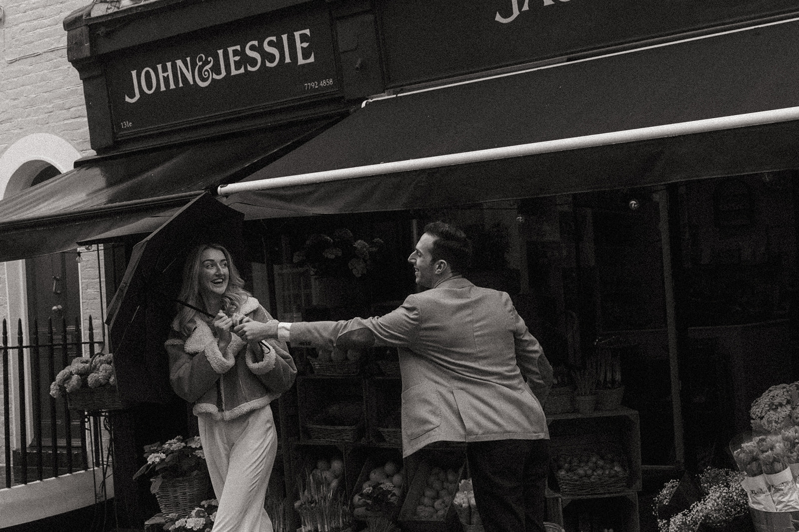A man reaches for the umbrella from a woman, they are both laughing, shot by London engagement photographer Elsie Goodman.