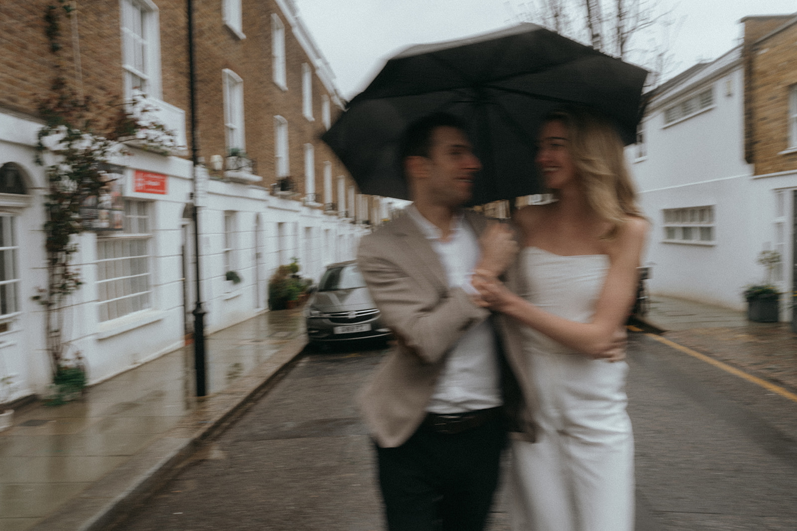 Blurry action shot of a man and woman walking under an umbrella, smiling at each other, shot by London engagement photographer Elsie Goodman.