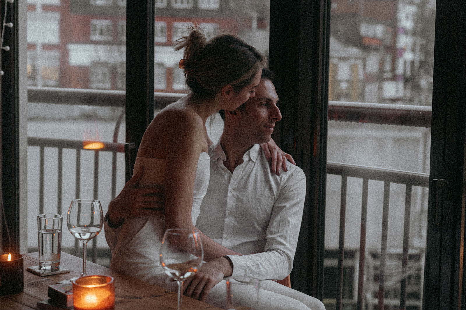 Woman sitting on a man's lap kissing the side of his forehead as he looks out the window at a rainy London street, shot by London engagement photographer Elsie Goodman.