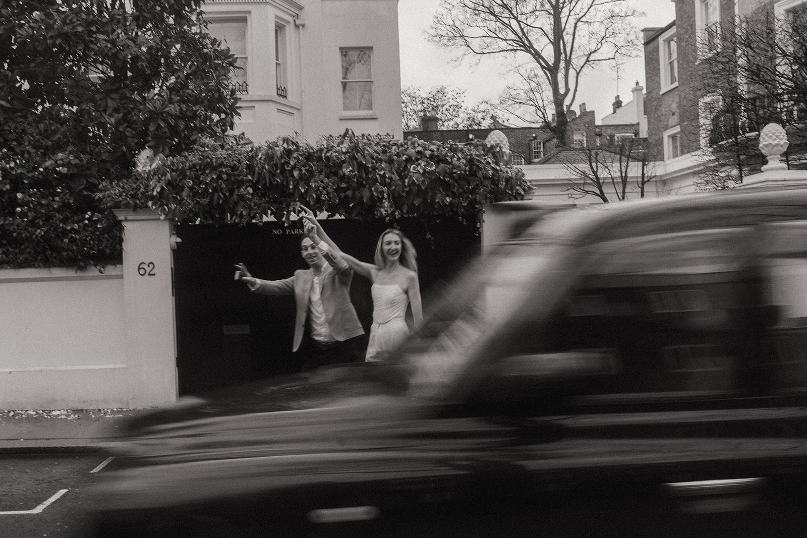 Man and woman smiling with their hands in the air as a passing car speeds through the shot, shot by London engagement photographer Elsie Goodman.