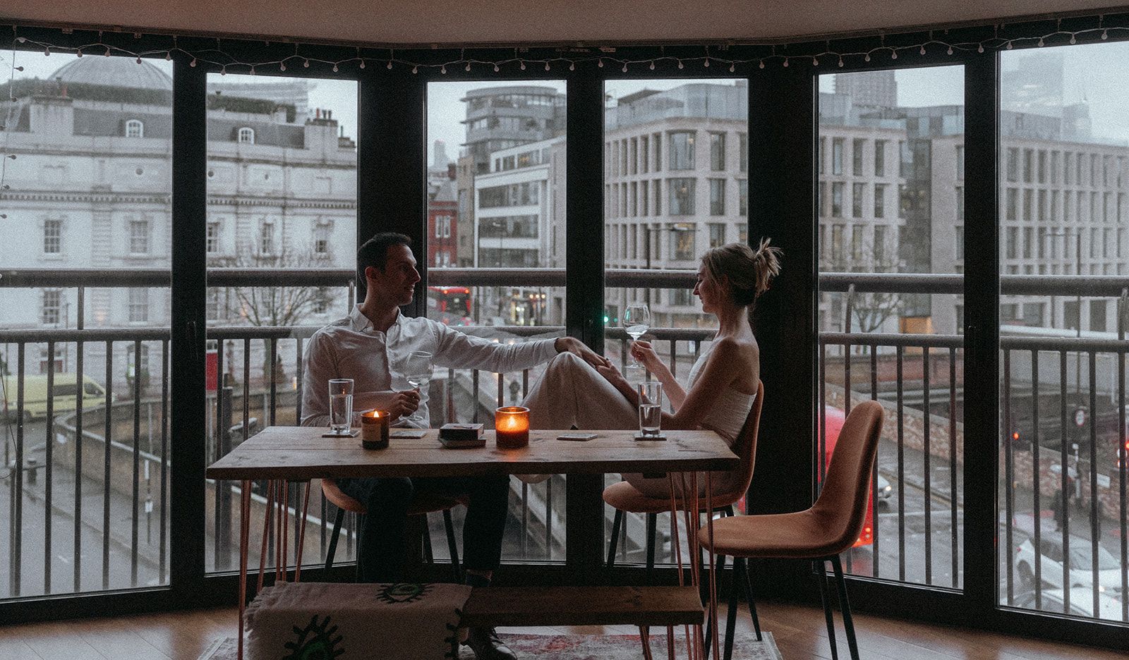 Wide angle shot of a man and women sitting at a table with a stormy London street behind them. They are both looking at each other, holding hands over her knees, shot by London engagement photographer Elsie Goodman.