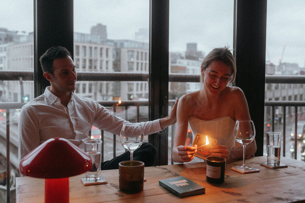 Man and women sit at a table with a dark London street visible through the windows behind them. She is lighting a candle with a match, he has his arm resting on her back, they are both smiling, shot by London engagement photographer Elsie Goodman.