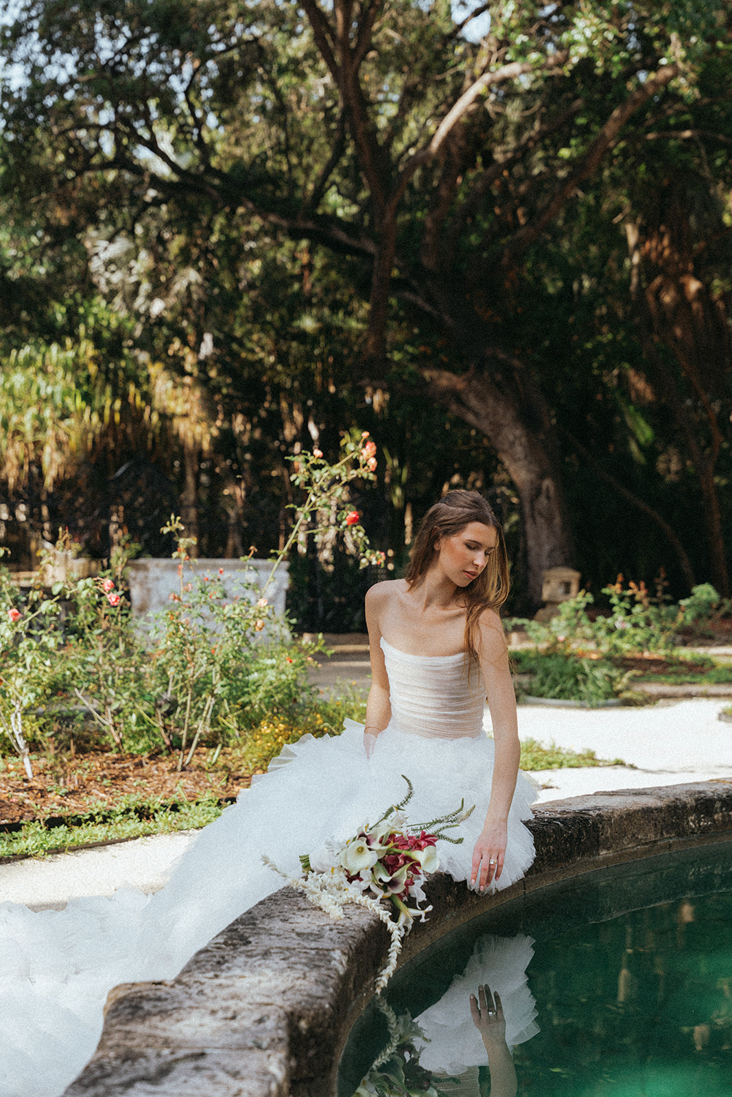 Viscaya Bride sitting on a concrete barrier looking down into a pool of water by Vizcaya Wedding Photographer Elsie Goodman.
