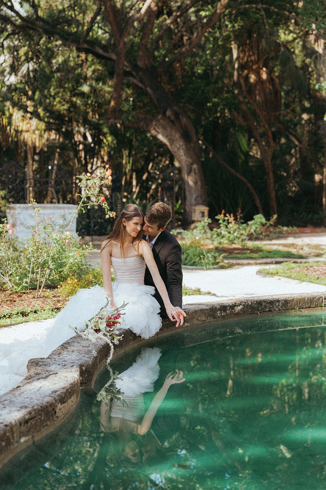 Viscaya Groom sits behind his bride on the edge of a fountain leaning into each other by Vizcaya Wedding Photographer Elsie Goodman.