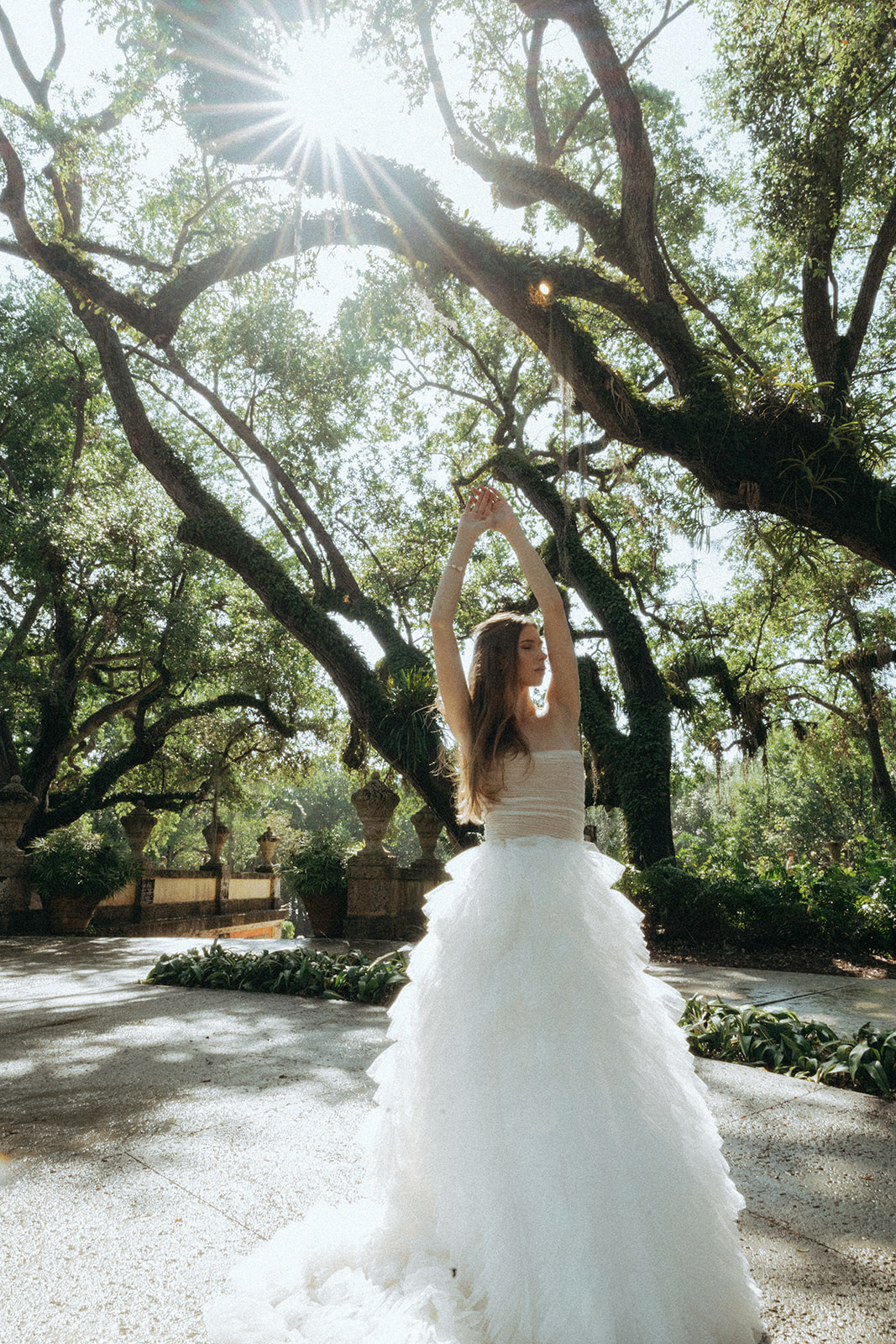 Viscaya Bride standing in the light, under old trees with her hands stretched overhead by Vizcaya Wedding Photographer Elsie Goodman.