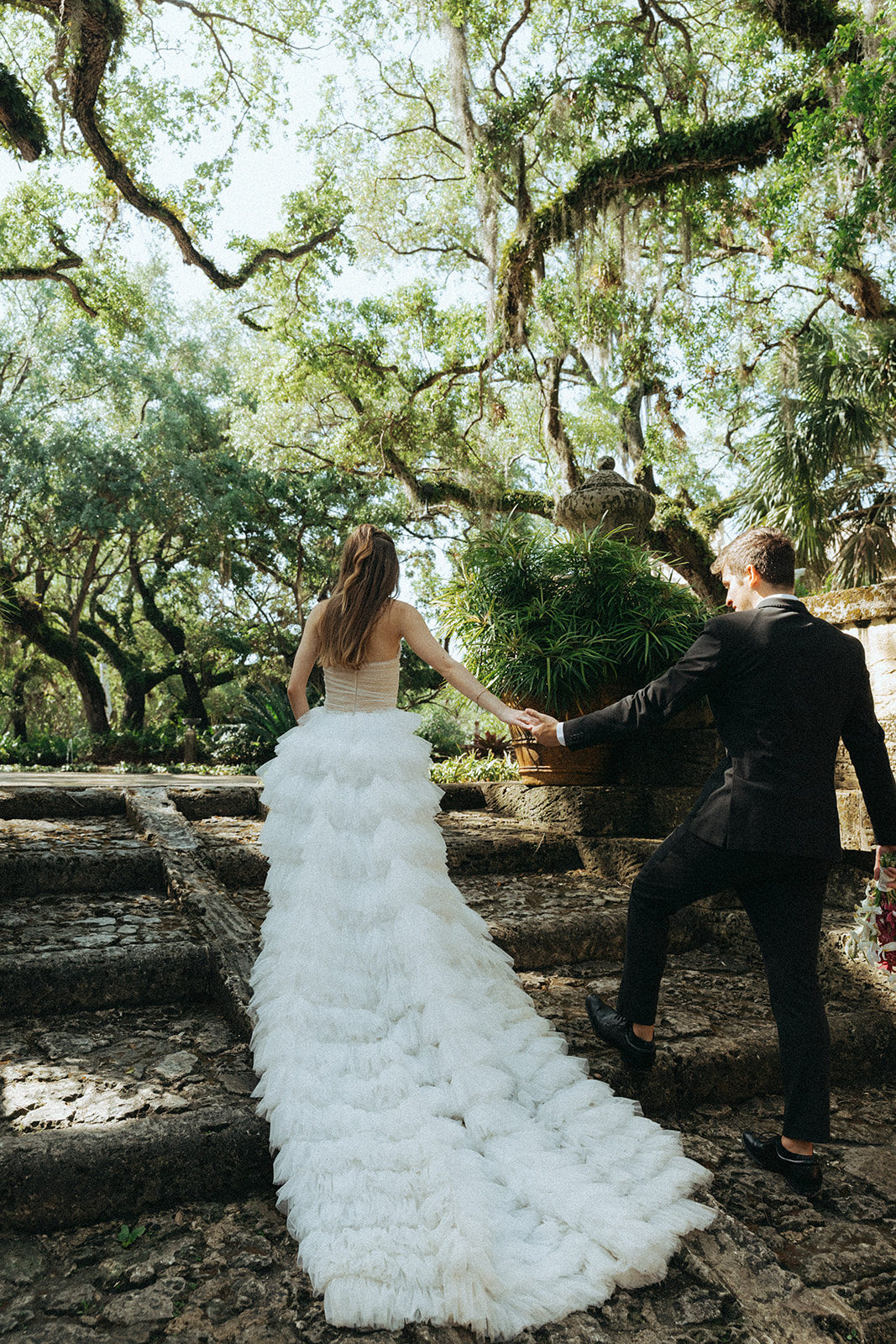 Viscaya Bride walking up large steps, showing off her long dress train, her groom holds her hand to help steady her by Vizcaya Wedding Photographer Elsie Goodman.