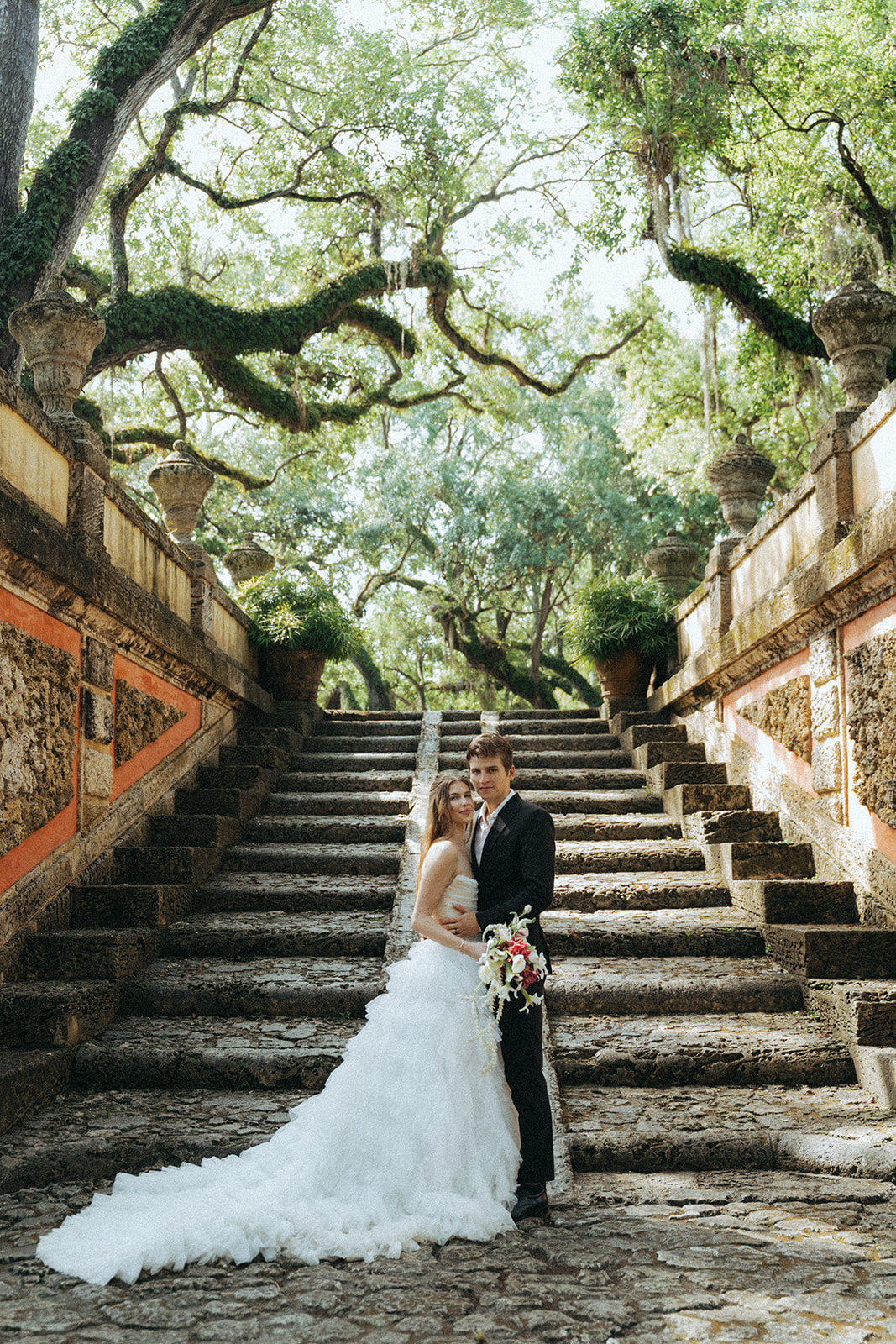 Viscaya Bride and groom pose looking at the camera with a big outdoor staircase and mossy trees by Vizcaya Wedding Photographer Elsie Goodman.