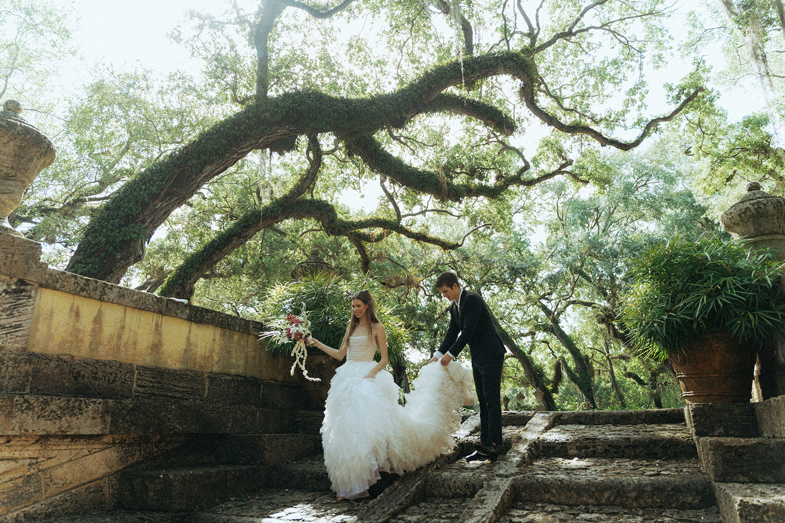 Viscaya bride walks down a staircase holding her bouquet, her groom helps unfold her train behind her by Vizcaya Wedding Photographer Elsie Goodman.