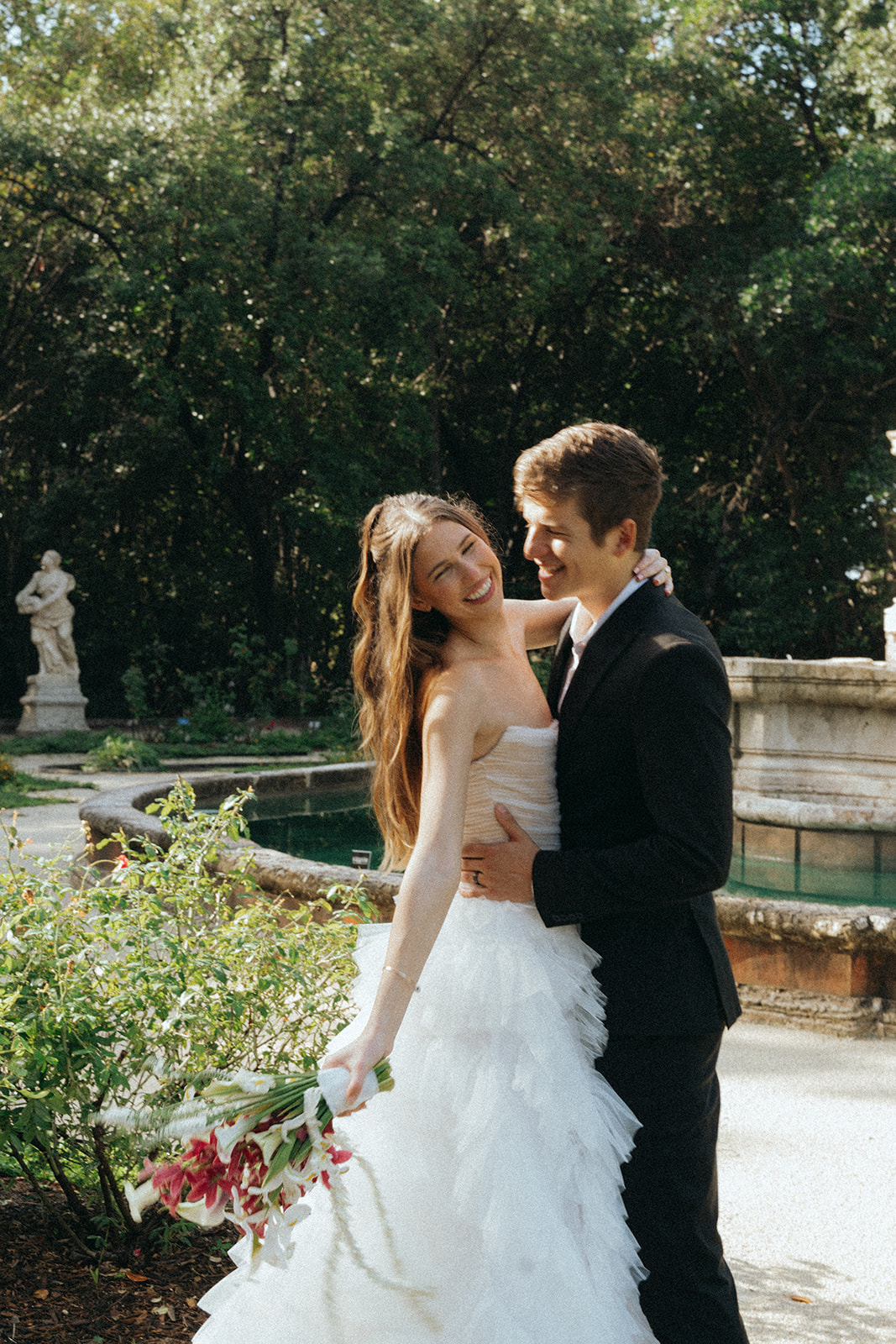 Viscaya Groom has his arms wrapped around her, she's smiling looking at the camera with her bouquet in one hand by Vizcaya Wedding Photographer Elsie Goodman.