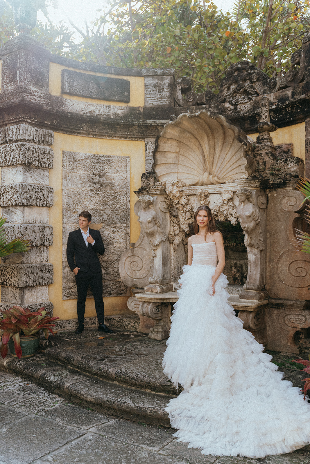 Viscaya Bride stands looking at the camera, her dress flowing down two steps, the groom stands further behind with his back against the wall with one hand on his suit jacket and one in his pocket by Vizcaya Wedding Photographer Elsie Goodman.