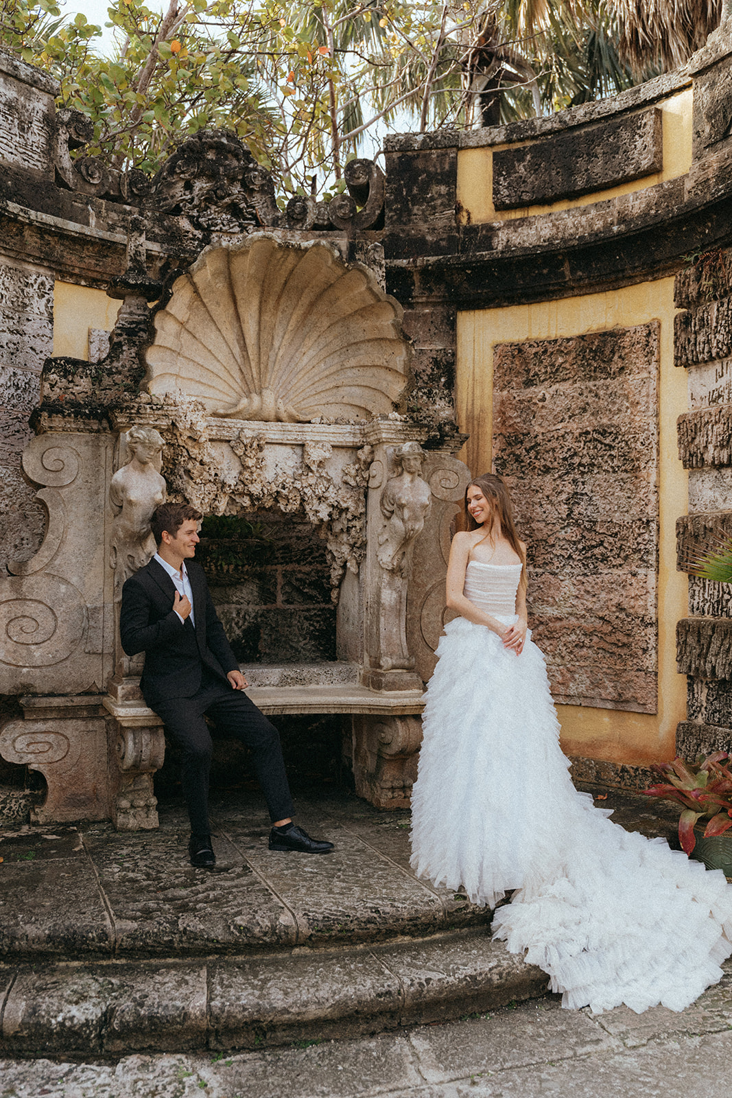 Viscaya Groom sits on a concrete garden bench looking at his bride by Vizcaya Wedding Photographer Elsie Goodman.