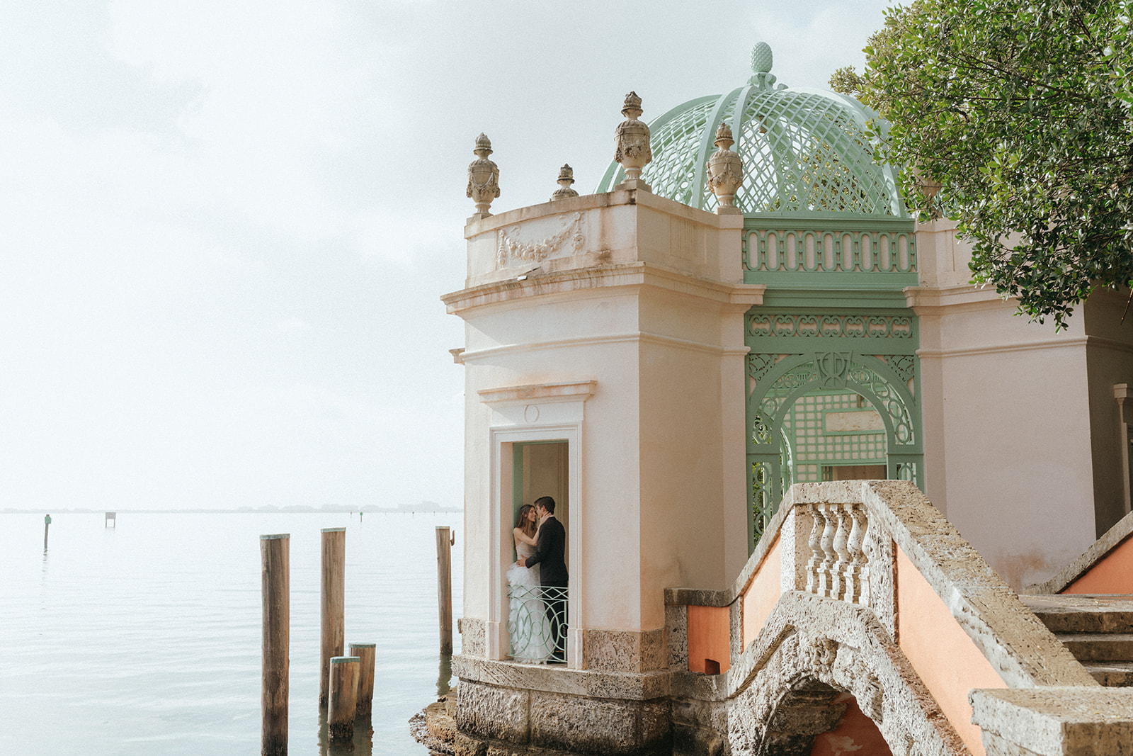 Viscaya Bride and groom kissing in a doorway of a gazebo, being photographed from further away so it shows the ocean by Vizcaya Wedding Photographer Elsie Goodman.