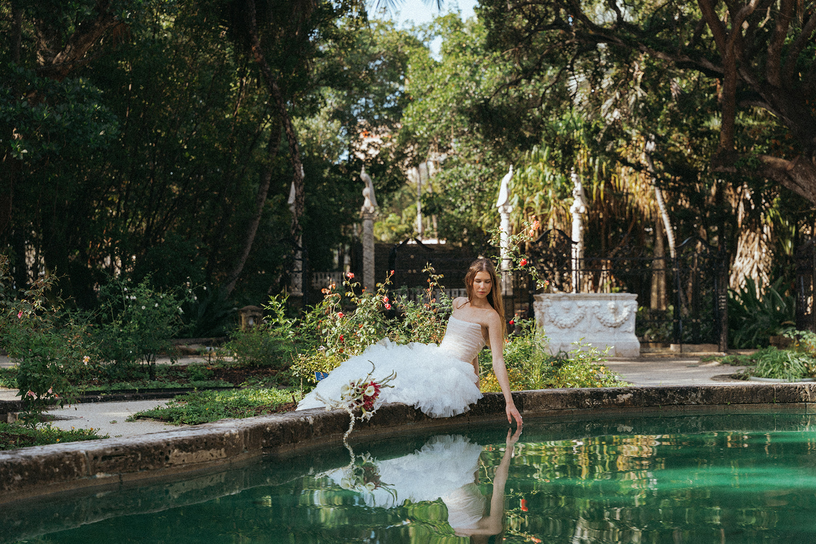 Viscaya Bride sitting on a concrete ledge, leaning over a pond. Her reflection in the water is clear by Vizcaya Wedding Photographer Elsie Goodman.