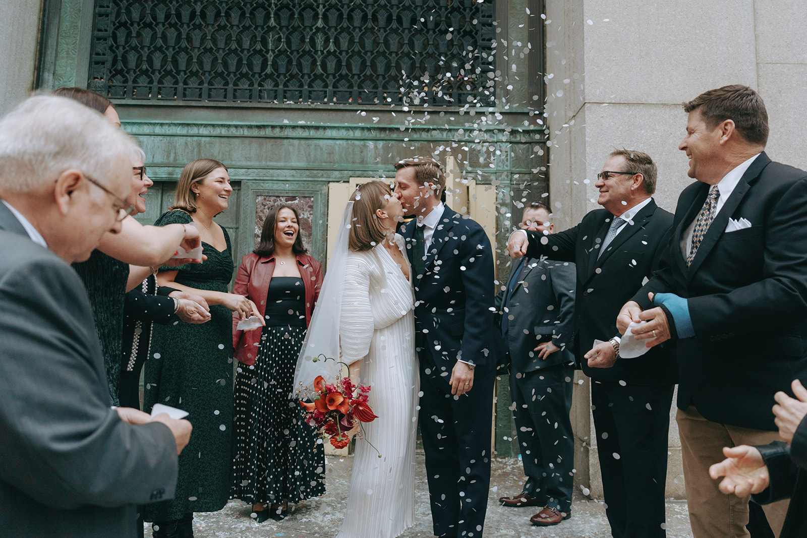 Rachel and Dave’s 2 day celebration Bride and groom kiss with friends and family on either side throwing confetti in front of city hall by NYC city hall wedding photographer Elsie Goodman.